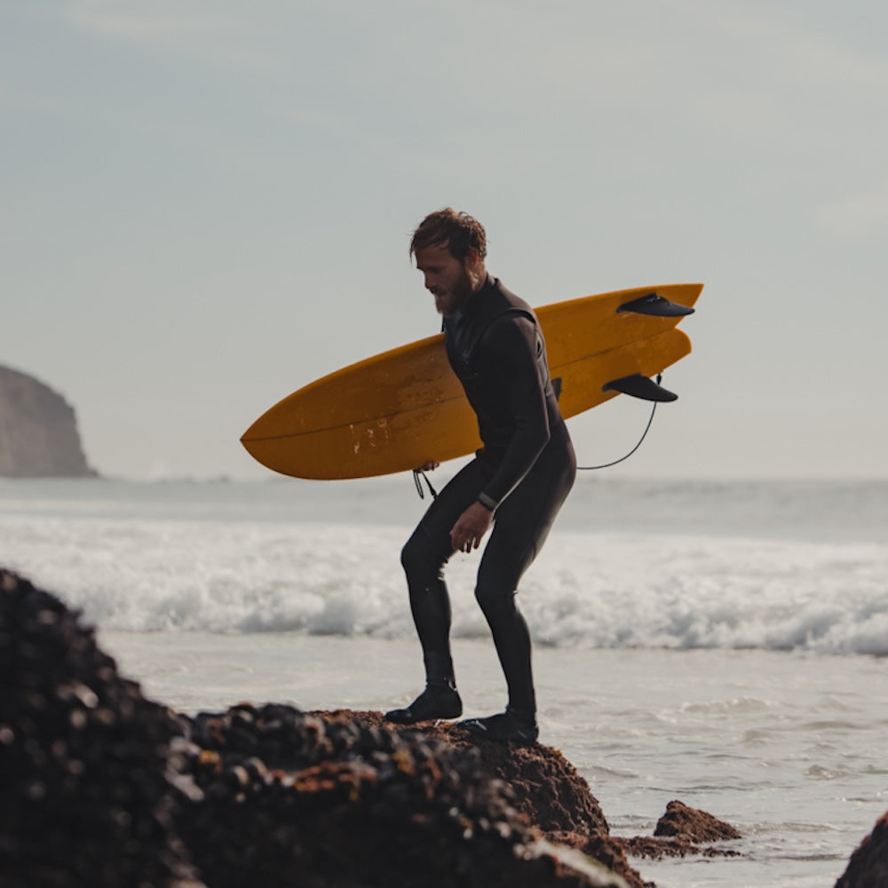 Man in a wetsuit holding a yellow surfboard on a rocky beach.