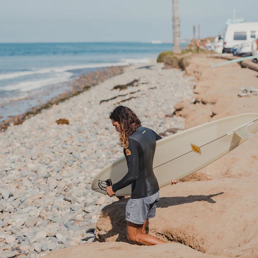 Person in a wetsuit holding a surfboard on a pebbly beach with ocean and boat in the background