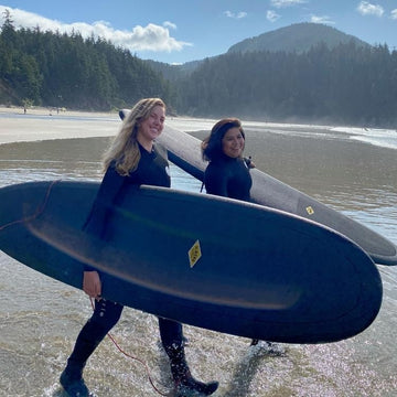 Two people in wetsuits carrying a large blue surfboard on a beach with trees and mountains in the background.