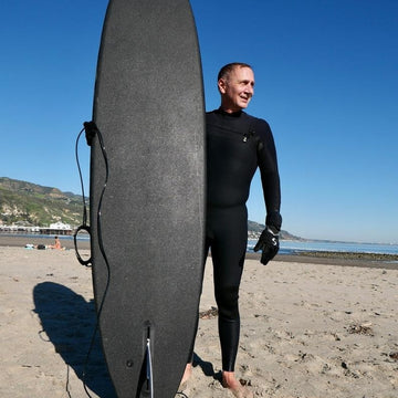 Man in a wetsuit holding a surfboard on a beach with a clear blue sky.