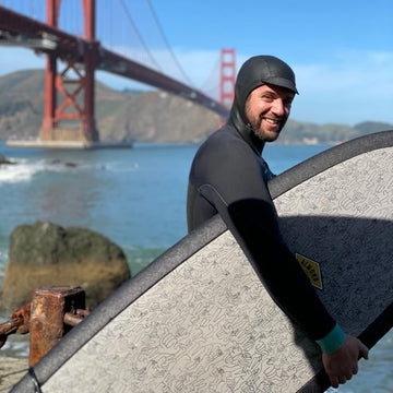 Man in a wetsuit holding a surfboard with the Golden Gate Bridge in the background