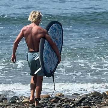 Man with a surfboard walking on a rocky beach towards the ocean
