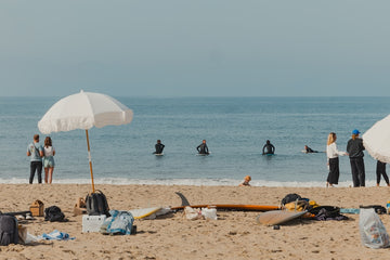 Beach scene with people, surfboards, and an umbrella on a sandy shore.