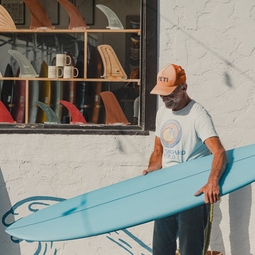 Man holding a blue surfboard in front of a store with surfboards and mugs displayed.