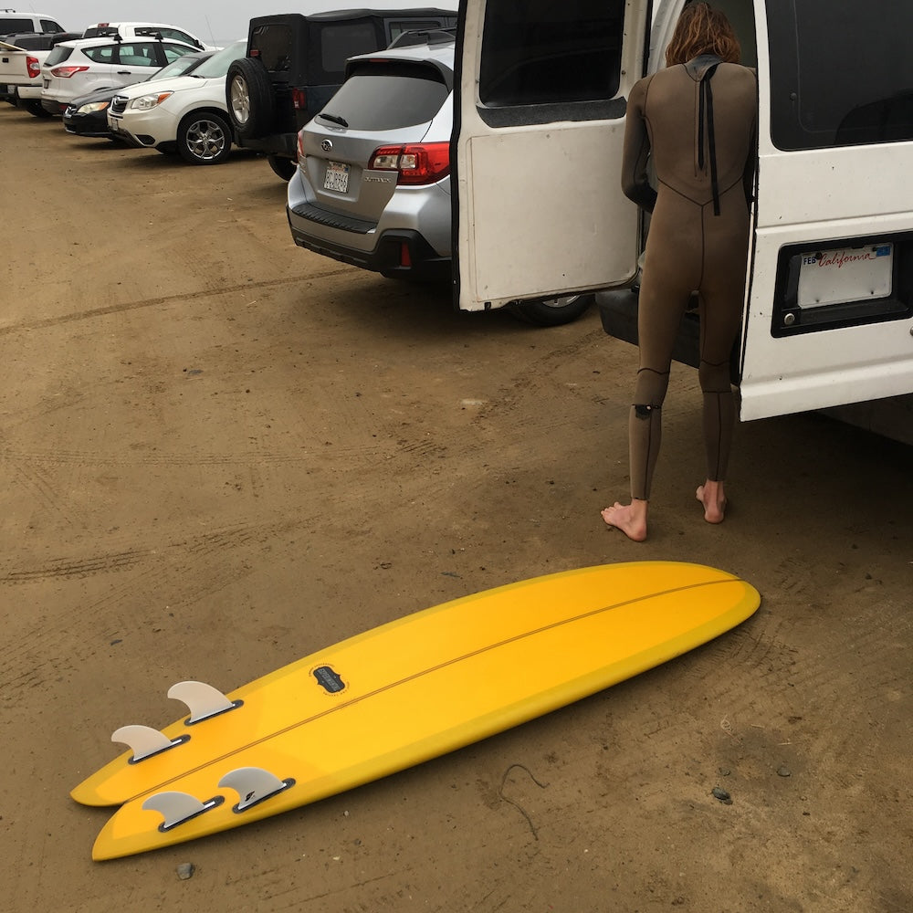Person in a wetsuit getting out of a van with a yellow surfboard on the ground.