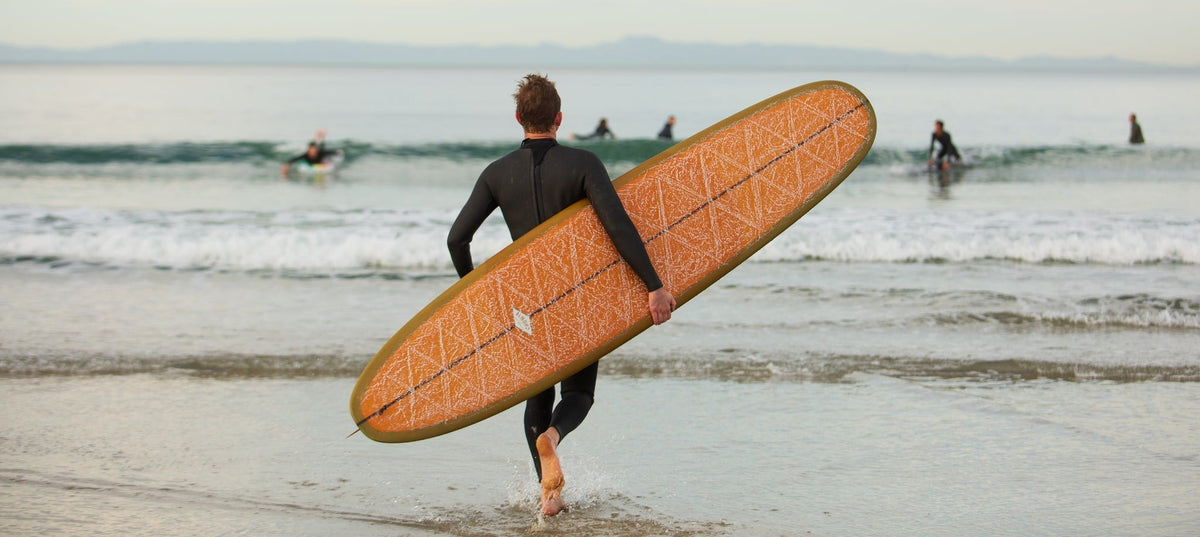 Person in a wetsuit carrying a longboard surfboard on a beach with other surfers in the background.