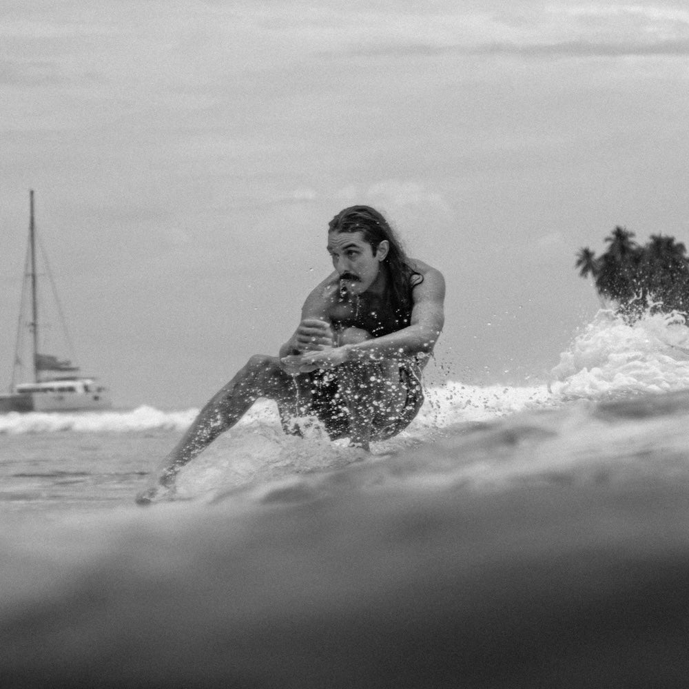 Person surfing on a wave with a sailboat in the background
