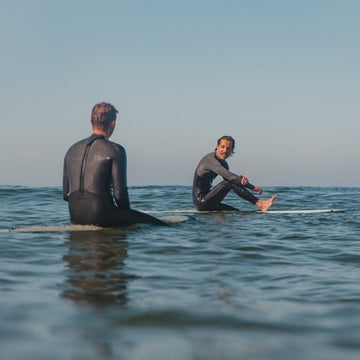 Two people in wetsuits sitting on surfboards in the ocean with a clear sky.