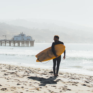 Person in a wetsuit holding a yellow surfboard on a beach with a pier in the background