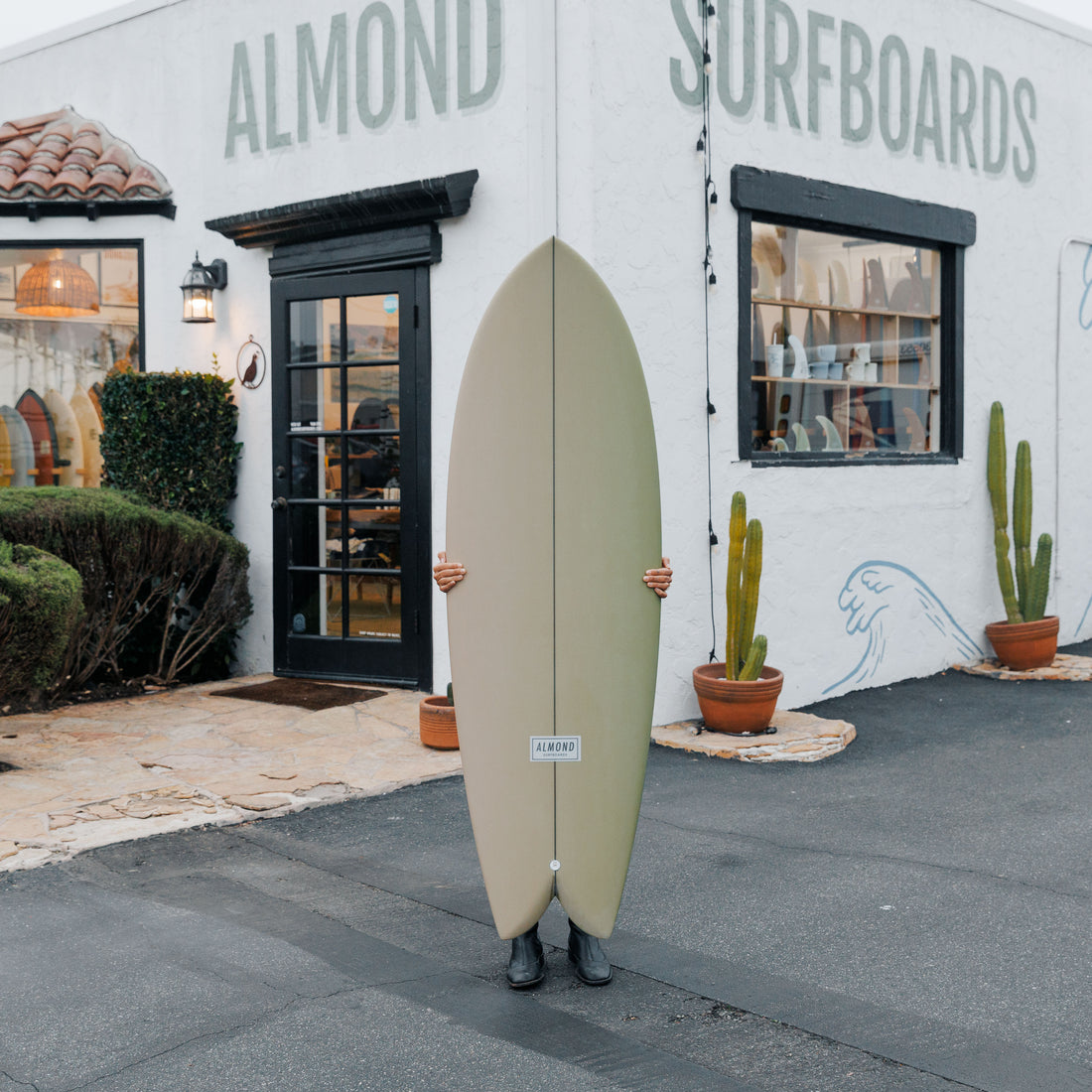 Person holding a surfboard in front of Almond Surfboards store