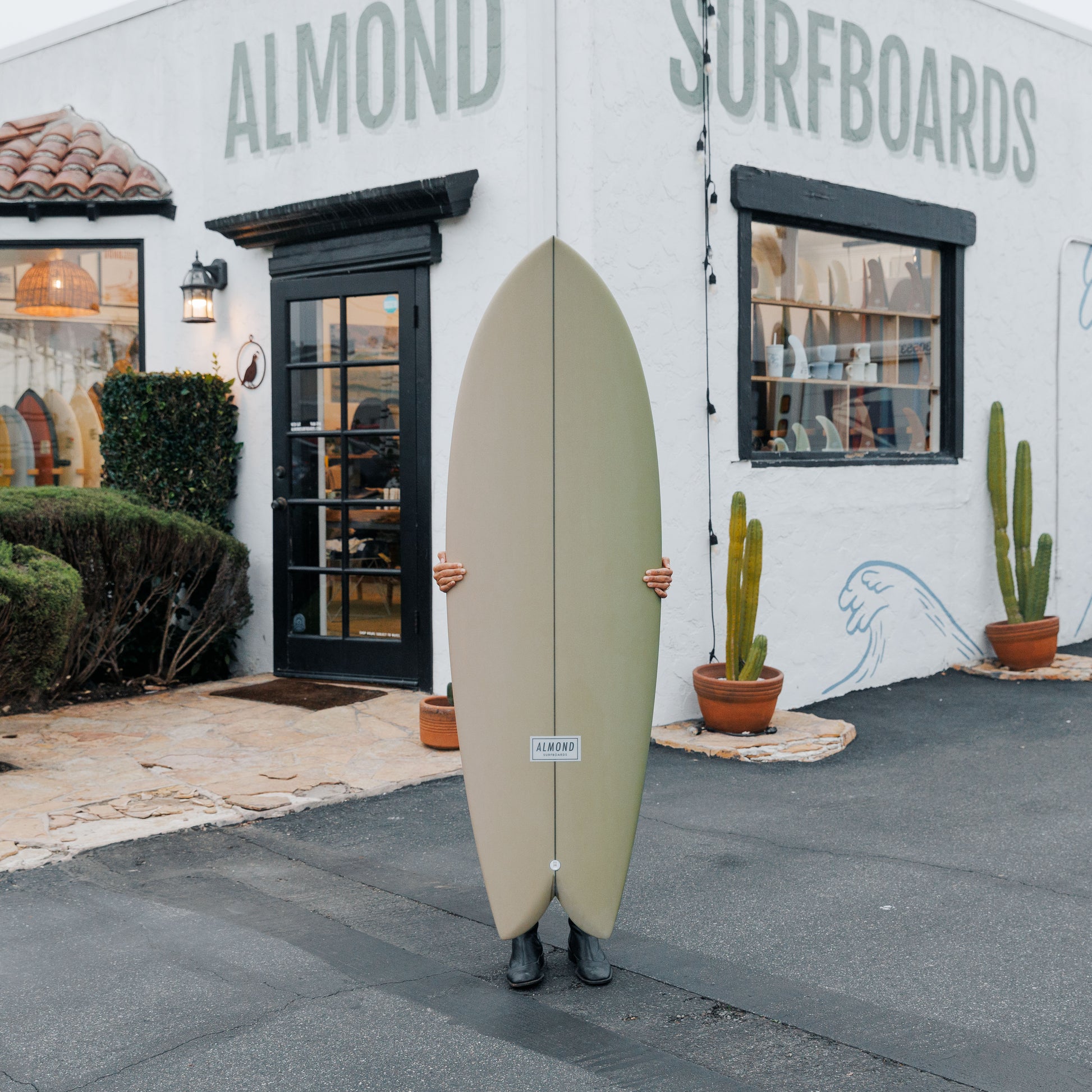 Person holding a surfboard in front of Almond Surfboards store