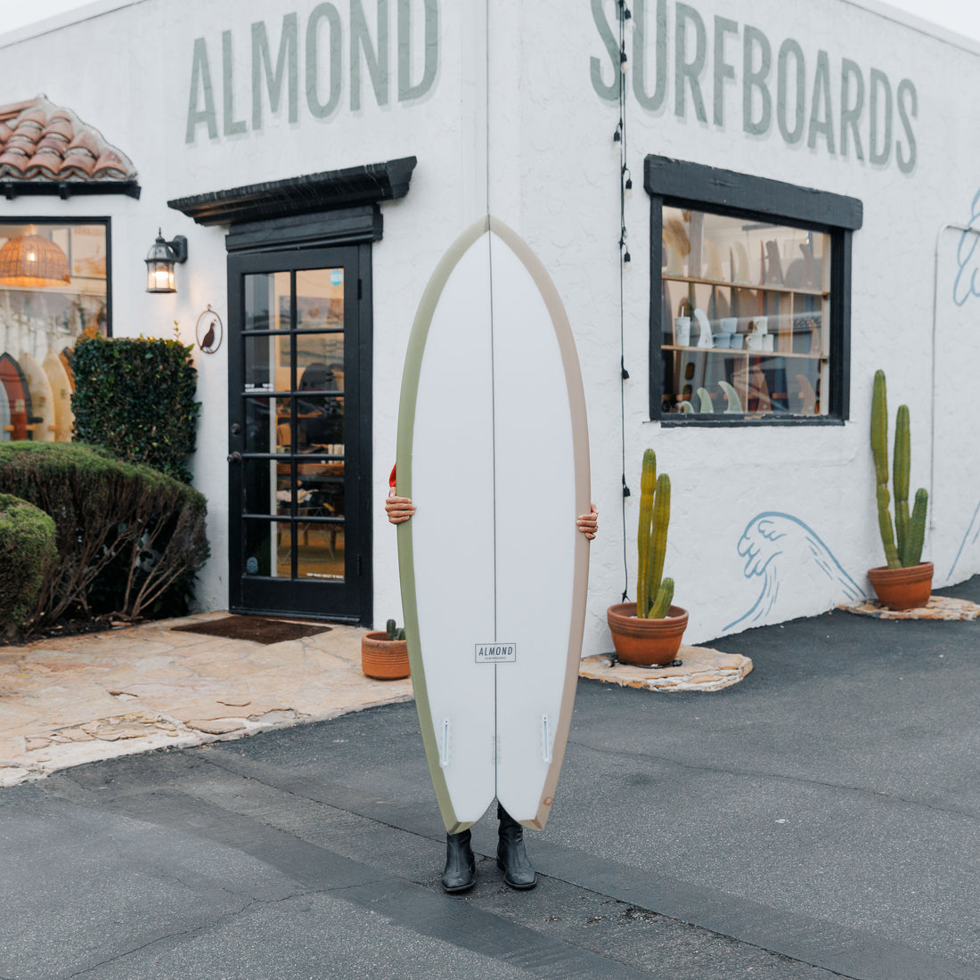 Person holding a surfboard in front of Almond Surfboards store