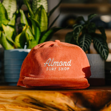 Orange cap with 'Almond Surf Shop' logo on a wooden surface with plants in the background