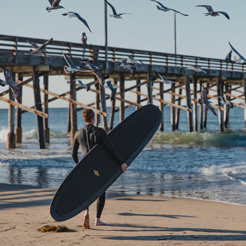 Person in a wetsuit holding a surfboard on a beach with a pier and birds in the background