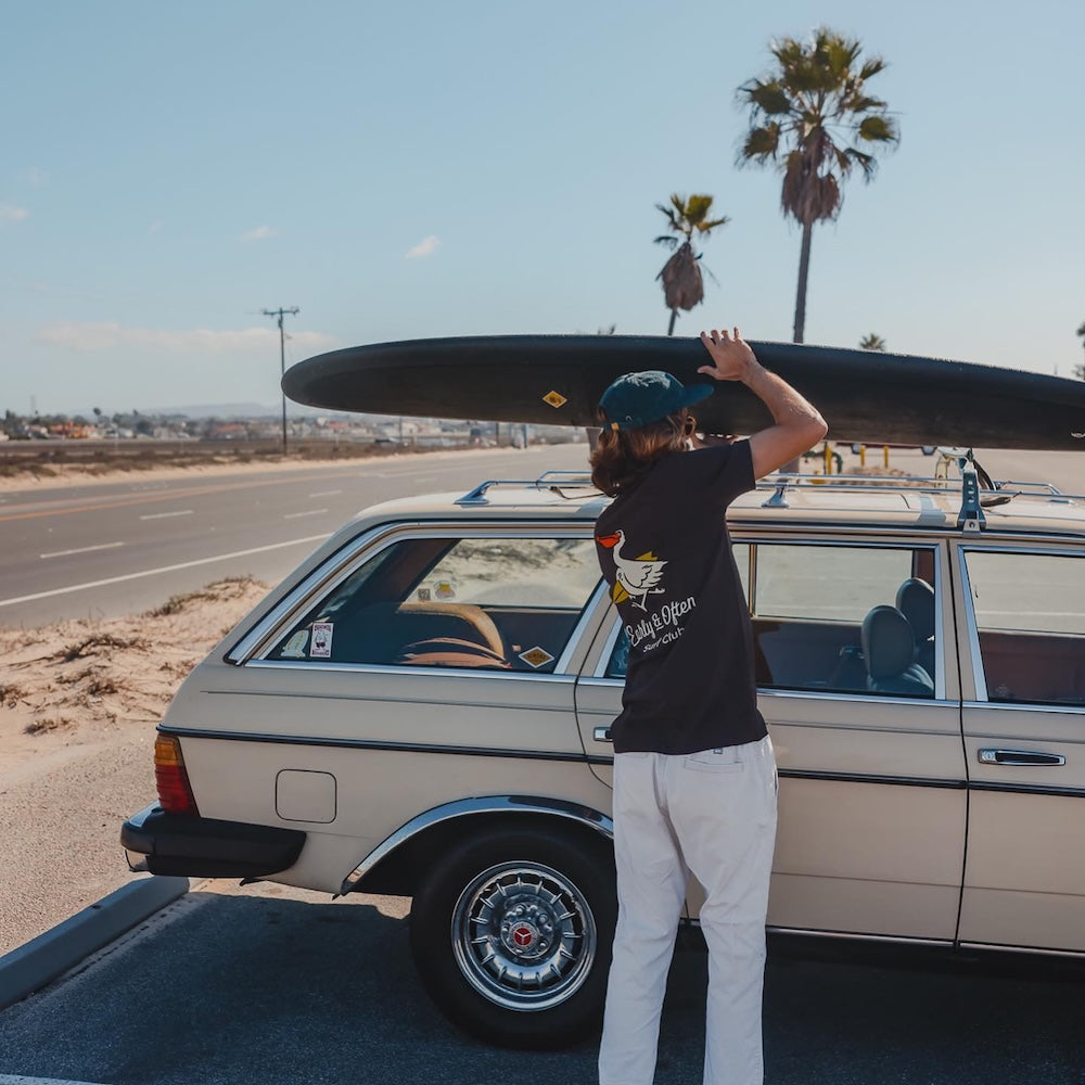 Person loading a surfboard onto a vintage car with palm trees in the background
