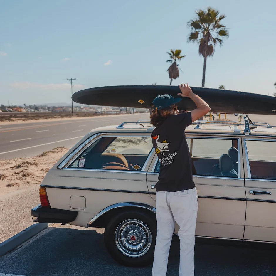 Person loading a surfboard onto a vintage car with palm trees in the background