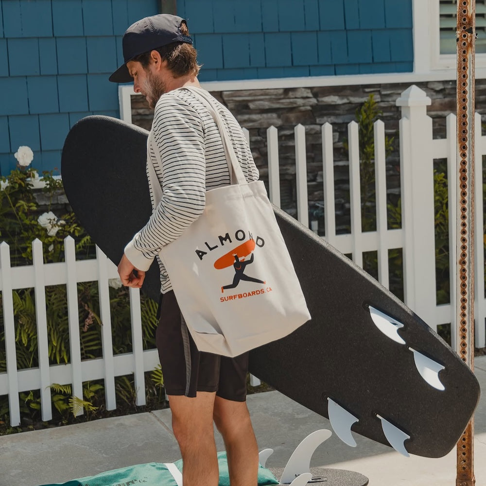 Man holding a surfboard with a tote bag featuring a surfboard design.