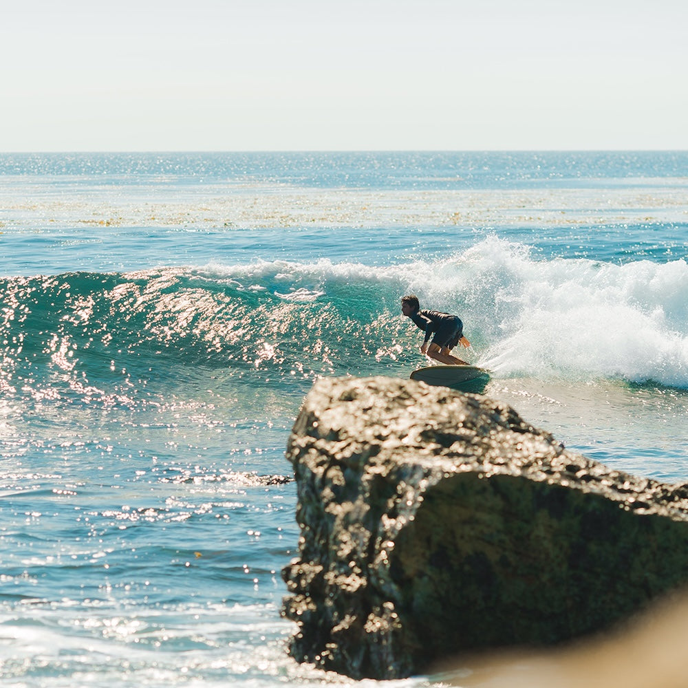 Person surfing on a wave near a rocky outcrop with clear blue water.