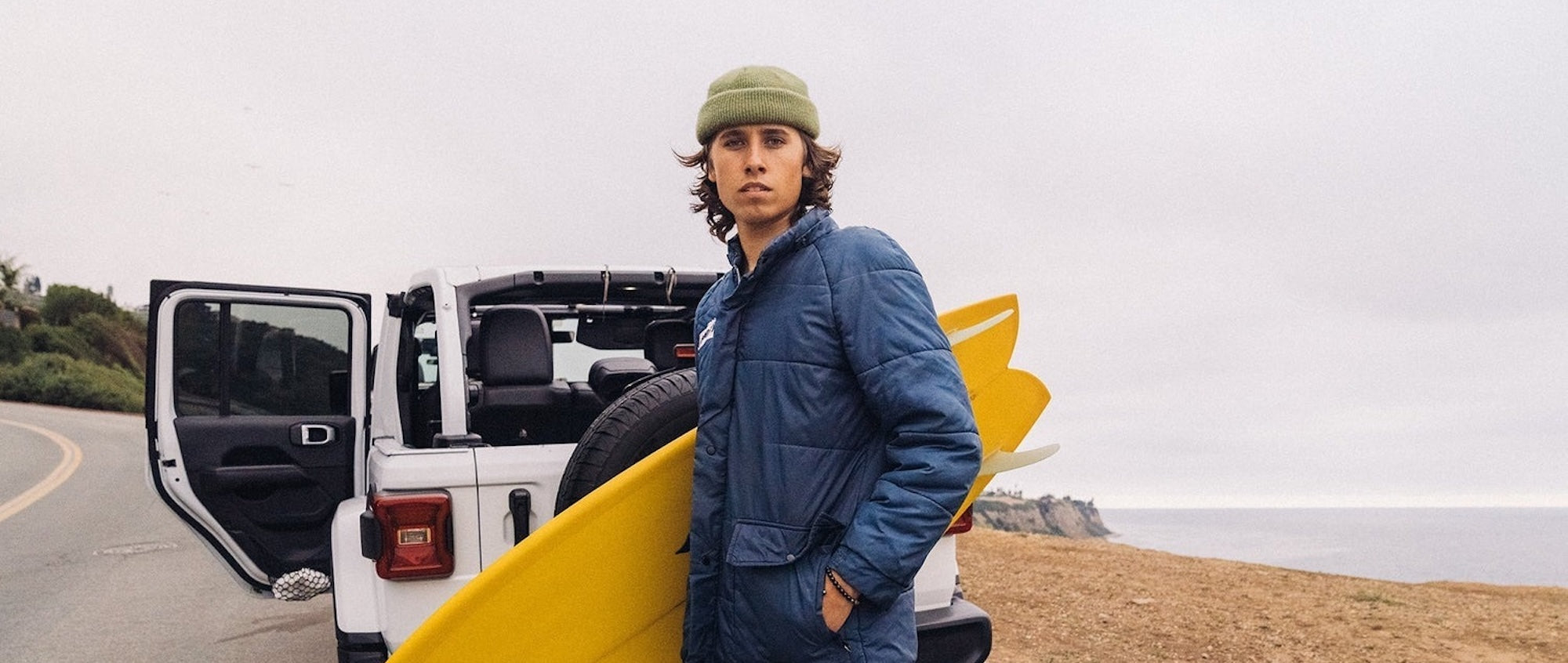 Person holding a yellow surfboard next to a white vehicle on a road.slider_item_9ybRfB