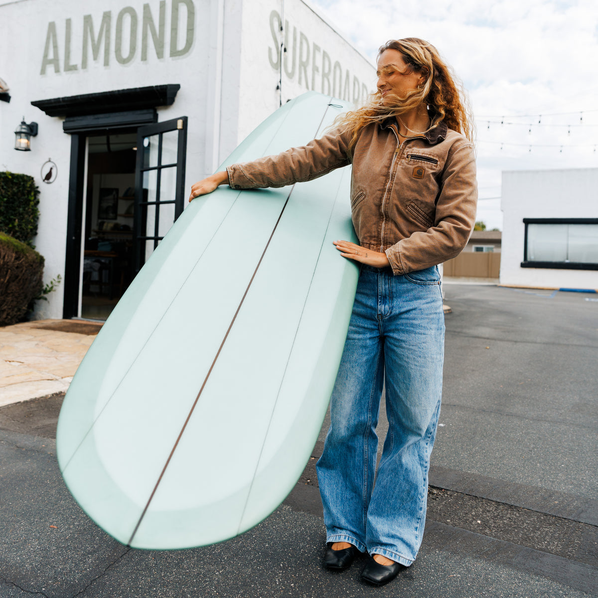 Person holding a surfboard in front of a building with 'Almond Surfboards' on it.
