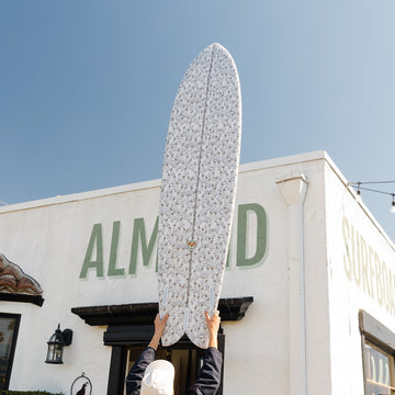 Person holding a surfboard in front of a building with 'ALM' branding.