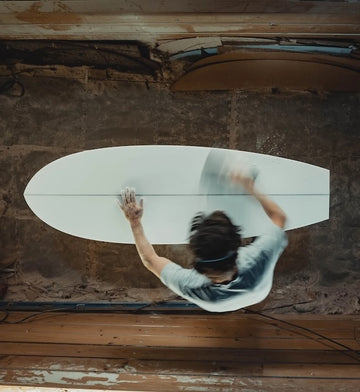 Person sanding a large white surfboard in a workshop.
